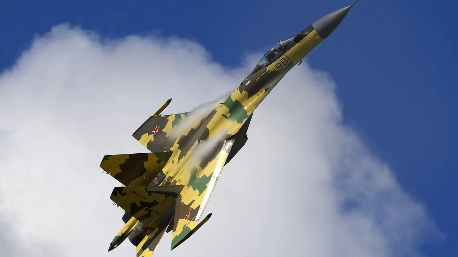 A Russian Air Force Sukhoi Su-35 in a green and yellow camouflage scheme climbing steeply against a blue sky and white cloud