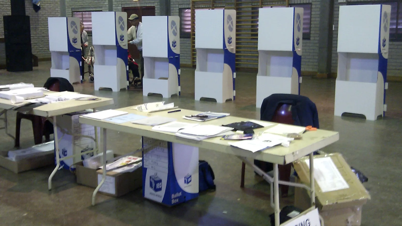 Inside a South African voting station: white IEC voting booths along a wall and tables with ballot boxes and papers