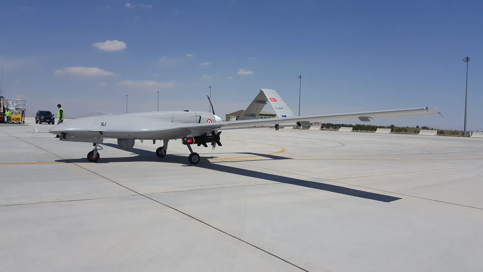 A white Bayraktar TB2 drone with a long, slender wing parked on a concrete apron under a blue sky
