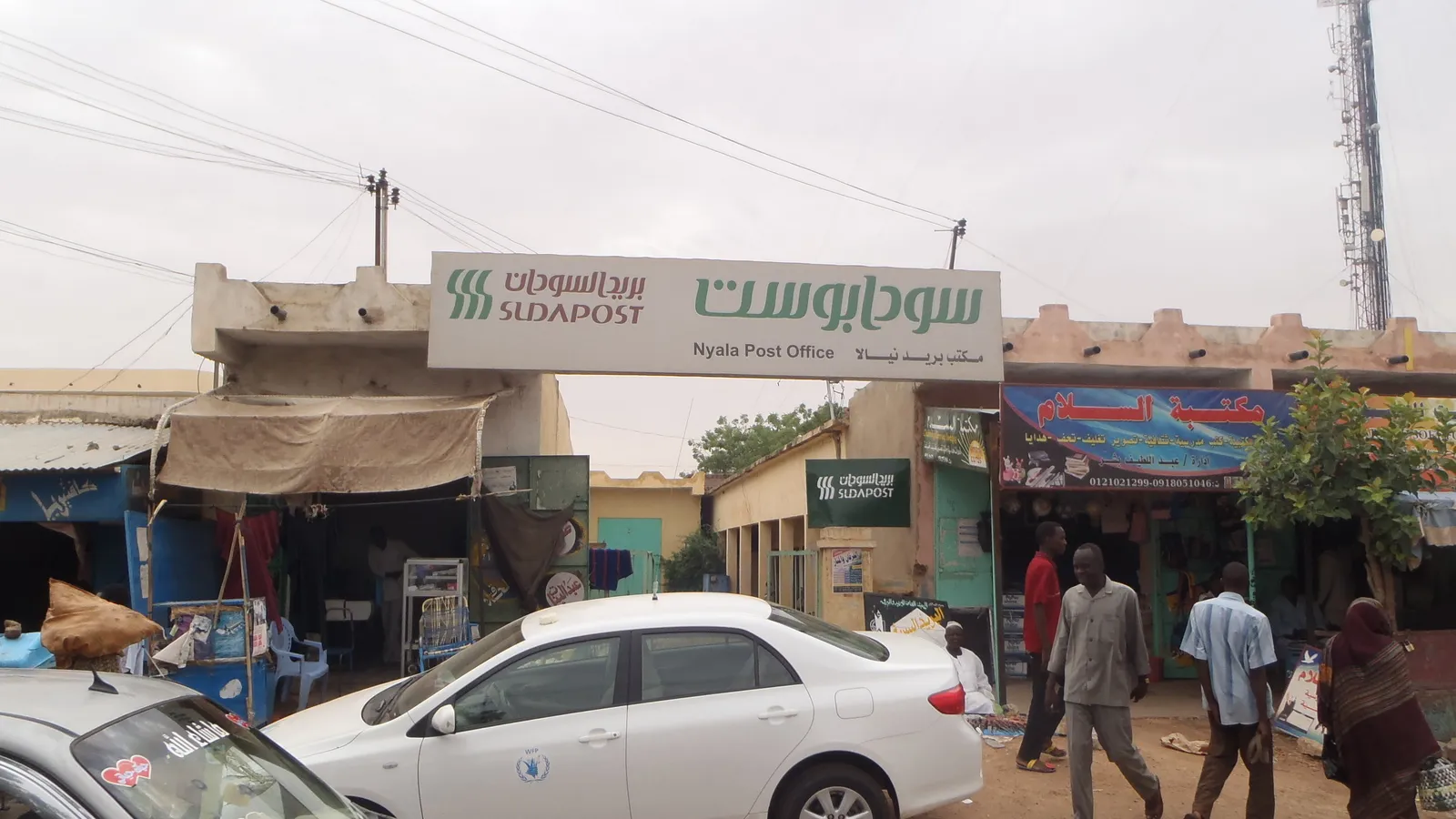 A street in Nyala with small shops, a white car and pedestrians in front of the Sudan Post office building
